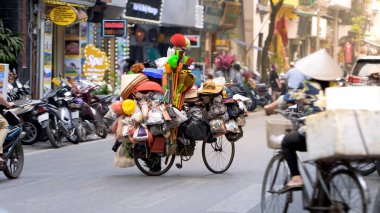 Hanoi, Vietnam-04 Nisan 2024: Hareketli şehir manzarasında, Vietnamlı bir sokak satıcısı temizlik ürünleri ve süpürge dolu bir bisikleti itip yol boyunca mobil bir kabine dönüştürüyor.