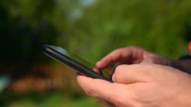 Parlak bir arka bahçede akıllı telefon tutan bir adam. Güneşli bir bahçede, açık havada telefon tutan erkek elleri. Doğal ışık, sakin bir ortamda rahat ve modern bir yaşam tarzını vurgular.