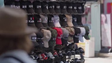 Güney Kore, Seul - 10 Temmuz 2025: Myeongdong, Seul 'da bir sokak tezgahında raflarda dizilmiş farklı renkli şapkaların yavaş çekimleri ve arka planda müşteriler var.