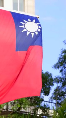 Dikey Tayvan bayrakları zarif bir şekilde bir direğin üzerinde dalgalanıyor. Arkadaki ağaçlara ve modern bir binaya karşı dizilmiş. Tayvan gururunu ve mirasını simgeliyor..