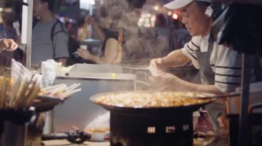 Güney Kore, Seul -08 Temmuz 2025: Myeongdong gece pazarında genç bayana sıcak bir tteokbokki sunan Koreli sokak yemekleri satıcısı yavaş hareket ediyor. Geleneksel yemek, gece ticareti ve yerel kültür..