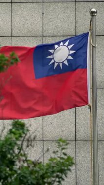 Arka planda modern bir bina bulunan ve ağaç dalları arasından görülen Tayvan bayrağının dikey yavaş hareketi. Sahne, ulusal gururu ve doğa ile şehir hayatının bütünleşmesini yakalıyor..