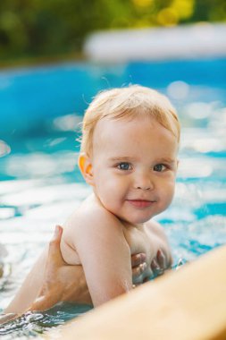 Kızıl saçlı küçük bir çocuğun portresi bahçede açık havuzda el desteği ile yıkanır, bebek suda oynar, aile yaz tatili yapar.