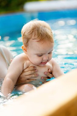 Kızıl saçlı küçük bir çocuğun portresi bahçede açık havuzda el desteği ile yıkanır, bebek suda oynar, yaz tatili yapar.