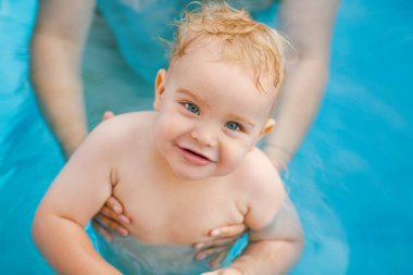 Kızıl saçlı küçük bir çocuğun portresi bahçede açık havuzda el desteği ile yıkanır, bebek suda oynar, yaz tatili yapar.