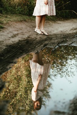 Kırsal yolda kirli su birikintisinde genç ve romantik bir kadının yansıması. Atmosferik bir resimde yaz elbisesi giymiş bir kızın yansıması.