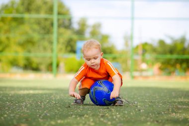 Futbol sahasında oturan iki yaşında bir çocuk, top oynayan küçük sarışın bir çocuk, aktif yaz tatili ve spor konsepti.