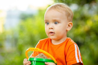 Dışarıdaki oyun parkındaki kum havuzunda oynayan sarışın bir çocuk. Bir kova dolusu kumu olan bir bebek portalı, yaz çocuğu eğlence aktivitesi.