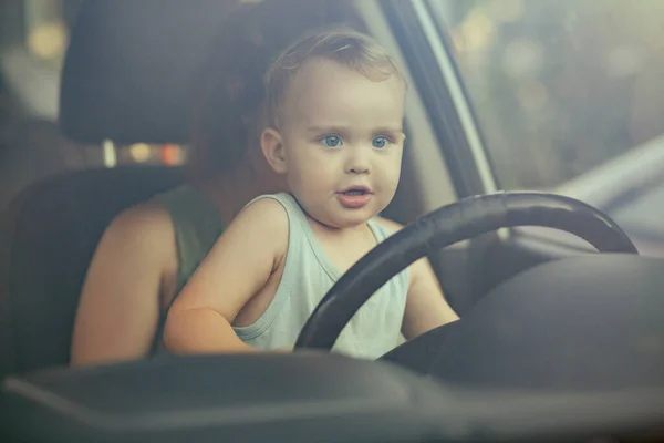child sitting on mother's knees in the car and turning steering wheel, little cute blond boy drive car for fun, family concept