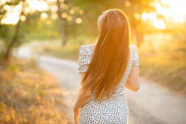 Günbatımında orman ekiminde yürüyen, yol üzerindeki sıcak güneş ışığıyla aydınlatılan, doğa kavramında rahatlayan genç, romantik bir kadın.