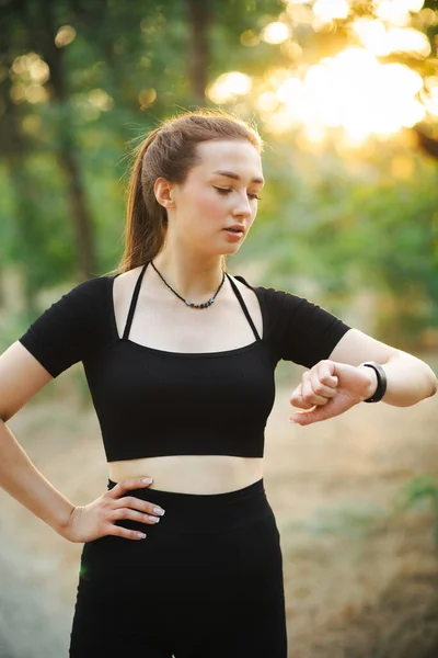 Young runner woman looking at the smart clock checking time and pulse in forest park. Girl training at sunset on nature, health and sport concept