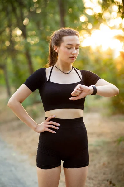 Young runner woman looking at the smart clock checking time and pulse in forest park. Girl training at sunset on nature, health and sport concept