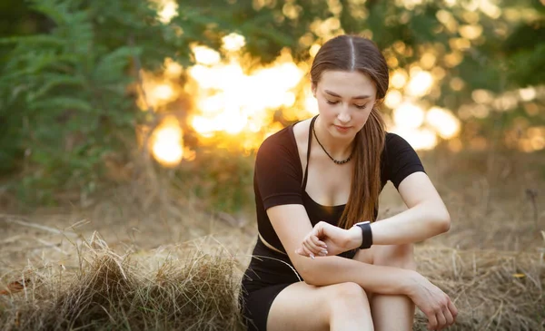 Young woman looking at the smart clock checking pulse sitting on the roadside in forest park. Girl training at sunset on nature, health and sport concept