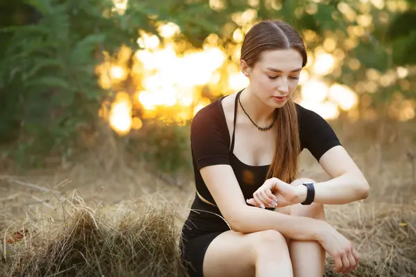 Young woman looking at the smart clock checking pulse sitting on the roadside in forest park. Girl training at sunset on nature, health and sport concept