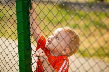 Yazın oyun bahçesinde demir çitlerin arkasında küçük sarışın çocuk, çocuk yaşam tarzı.
