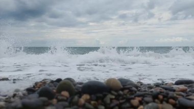 Okyanus dalgalarının çakıl taşı kaplı bir plajda yavaşça kırılıp bulutlu bir gökyüzünün altında çekildiği yavaş çekim videosu. Rahatlama sahnesi arka plan veya meditasyon kullanımı için mükemmeldir.