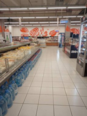 Geniş bir süpermarket koridorunun bulanık ve bulanık görüntüsü. Perspektif görünümü, ürün ve soğutulmuş kutularla dolu sıra sıra rafları gösteriyor, alışveriş veya perakende ticaret hissi iletiyor.