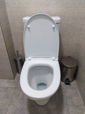 Temiz, modern bir banyoda, kapağı yukarıda beyaz bir klozet manzarası. Metal bir tuvalet fırçası tutacağı ve küçük metal bir çöp tenekesi gri fayanslı bir zeminde, birbirine uyumlu bir kiremit duvarın yanında.. 