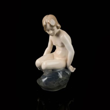 Statuette of a girl on a black background with reflection. porcelain antique figurine of a mermaid sitting on a stone