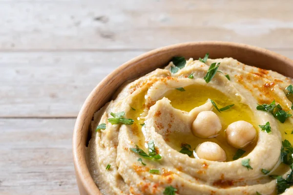 stock image Chickpea hummus in a wooden bowl garnished with parsley, paprika and olive oil on wooden table. Copy space