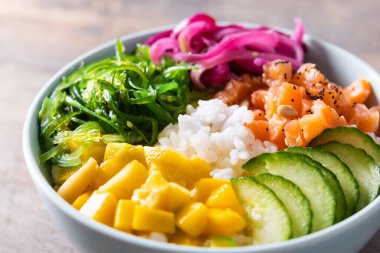 Poke bowl with rice, salmon,cucumber,mango,onion,wakame salad, poppy seeds ands sunflowers seeds on wooden background. Close up