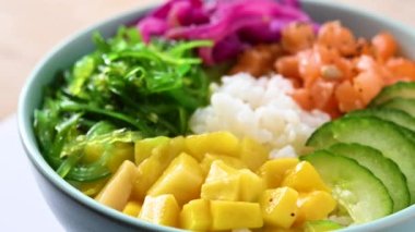 Poke bowl with rice, salmon,cucumber,mango,onion,wakame salad, poppy seeds ands sunflowers seeds circling on wooden table