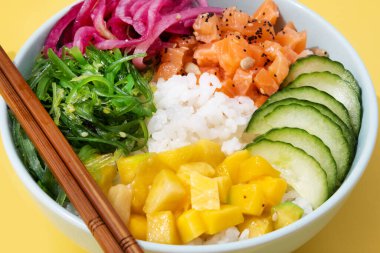 Poke bowl with rice, salmon,cucumber,mango,onion,wakame salad, poppy seeds ands sunflowers seeds on yellow background