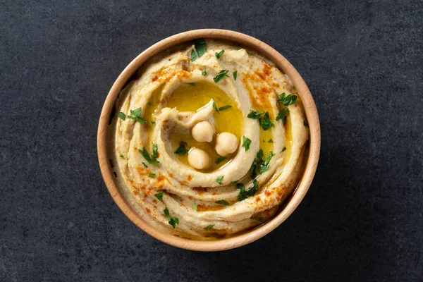 stock image Chickpea hummus in a wooden bowl garnished with parsley, paprika and olive oil on black slate background. Top view