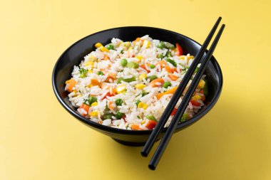 Sarı arka planda siyah bir kasede sebzeli beyaz pirinç.