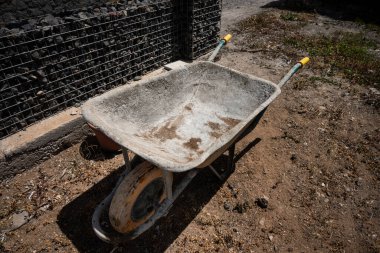 Tek ön tekerleği olan eski, yıpranmış bir el arabası tel ve taş çitin yanındaki kuru, tozlu toprakta duruyor. Metal gövdede ağır kullanım izleri var. Havzanın içinde pas ve kir birikti. Güneş ışığı gölgeler oluşturur, pürüzleri vurgular. 