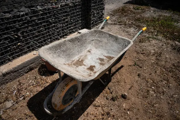 Tek ön tekerleği olan eski, yıpranmış bir el arabası tel ve taş çitin yanındaki kuru, tozlu toprakta duruyor. Metal gövdede ağır kullanım izleri var. Havzanın içinde pas ve kir birikti. Güneş ışığı gölgeler oluşturur, pürüzleri vurgular. 