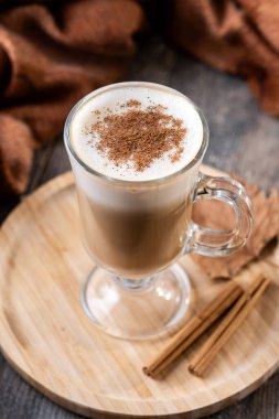 Tarçın tozuyla kaplı ılık akçaağaç latte, ahşap tepside cam bardakta servis edilir.