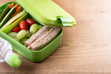 İçinde jambonlu peynirli sandviç, üzüm, vişneli domates ve sebze çubukları olan ahşap bir masanın üzerindeki yeşil beslenme çantası. Boşluğu kopyala