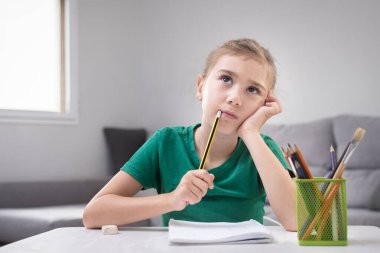 Yeşil tişört giyen genç bir kız elinde kalem ve defterle bir masada oturuyor.