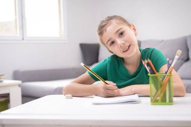 Yeşil tişörtlü mutlu genç kız elinde kalem ve defterle beyaz bir masada oturuyor. Ders çalışırken kameraya gülümsüyor..