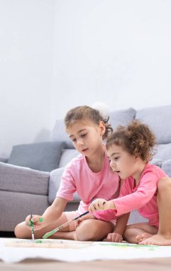 İki kız kardeş oturma odasında oturmuş, renkli boya ve fırçalarla birlikte büyük bir kağıt üzerinde resim yapıyorlar..