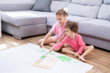 İki kız kardeş oturma odasında oturmuş, renkli boya ve fırçalarla birlikte büyük bir kağıt üzerinde resim yapıyorlar..