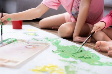 İki kız kardeş oturma odasında oturmuş, renkli boya ve fırçalarla birlikte büyük bir kağıt üzerinde resim yapıyorlar..