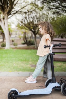Açık hava parkında üç tekerlekli scooterın üzerinde duran kıvırcık saçlı genç bir kızın yan görüntüsü. Günlük giysiler ve spor ayakkabılar giyen çocuk, doğada boş zamanların tadını çıkarıyor..