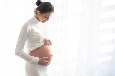 Beautiful brunette with a pregnant belly against the background of white window curtains with space for text.