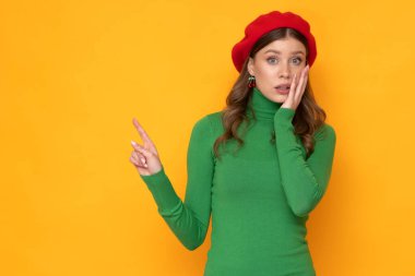 Shocked young woman with red beret and green sweater on isolated yellow background.