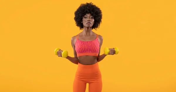 Dark skin female fitness trainer with afro hair and dumbbells on isolated orange background.