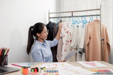 Tekstil aksesuarları ve girişimci tasarımcı çizimleriyle genç ve güzel Asyalı moda terzisi stüdyoda yaratıcı fikirler için dizüstü bilgisayarı olan parlak renklerle dolu..
