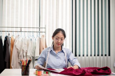 Tekstil aksesuarları ve girişimci tasarımcı çizimleriyle genç ve güzel Asyalı moda terzisi stüdyoda yaratıcı fikirler için dizüstü bilgisayarı olan parlak renklerle dolu..
