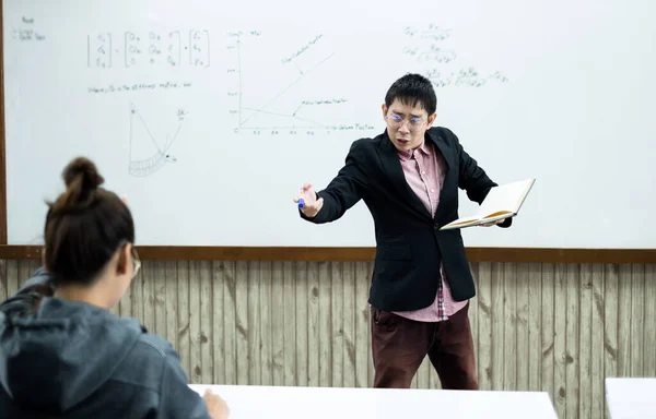 ホワイトボードの近くの教室では アジア系の男性教師が授業を作成し 授業のコラムで特別な講義を教えています 数学の先生は大学生への算術和を説明する — ストック写真