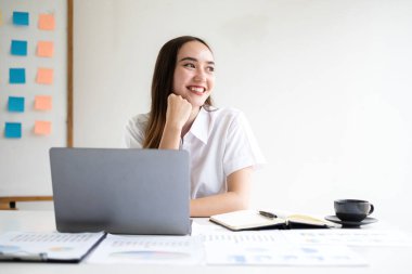 Asyalı iş kadını yeni bir sabah elinde kahveyle mutlu bir şekilde gülümsüyor. Ofiste çalışan boyunda bir kadını hafifçe yudumluyor..