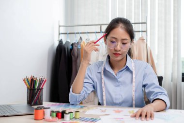 Tekstil aksesuarları ve girişimci tasarımcı çizimleriyle genç ve güzel Asyalı moda terzisi stüdyoda yaratıcı fikirler için dizüstü bilgisayarı olan parlak renklerle dolu..