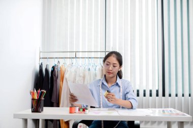 Tekstil aksesuarları ve girişimci tasarımcı çizimleriyle genç ve güzel Asyalı moda terzisi stüdyoda yaratıcı fikirler için dizüstü bilgisayarı olan parlak renklerle dolu..