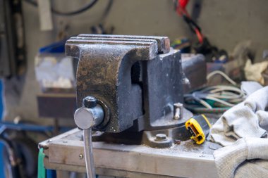 Sarı saplı siyah ve gümüş bir alet masanın üzerinde duruyor. Alet bir mengene ve bir atölye ayarında.