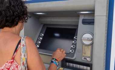 Bir kadın ATM makinesi kullanıyor. Kırmızı bir atlet ve mavi bir bilezik giyiyor.
